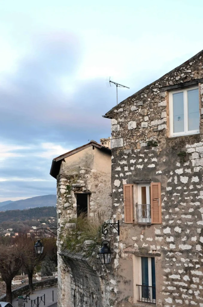 A close-up view of weathered stone buildings in a picturesque village, with soft pastel skies and mountainous terrain in the background in France