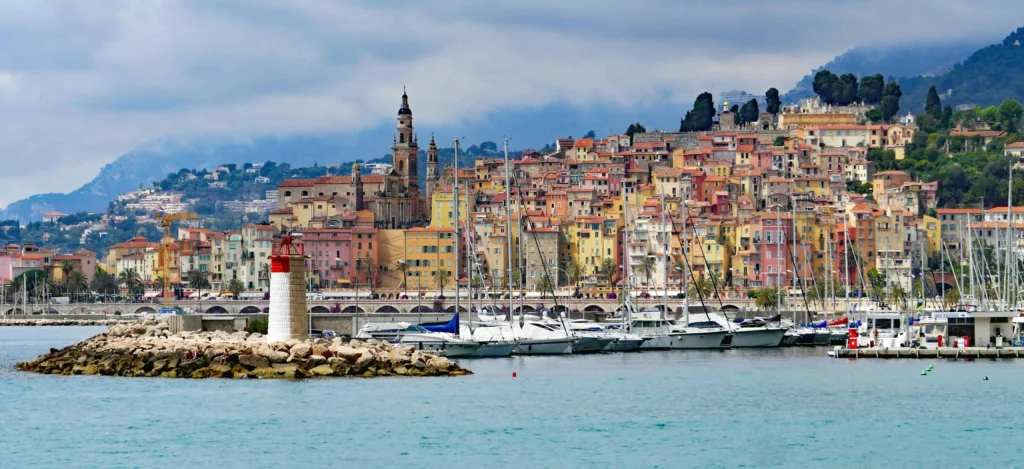 A picturesque harbor in the South of France, featuring colorful hillside houses, a red-and-white lighthouse, and moored yachts under a cloudy sky.