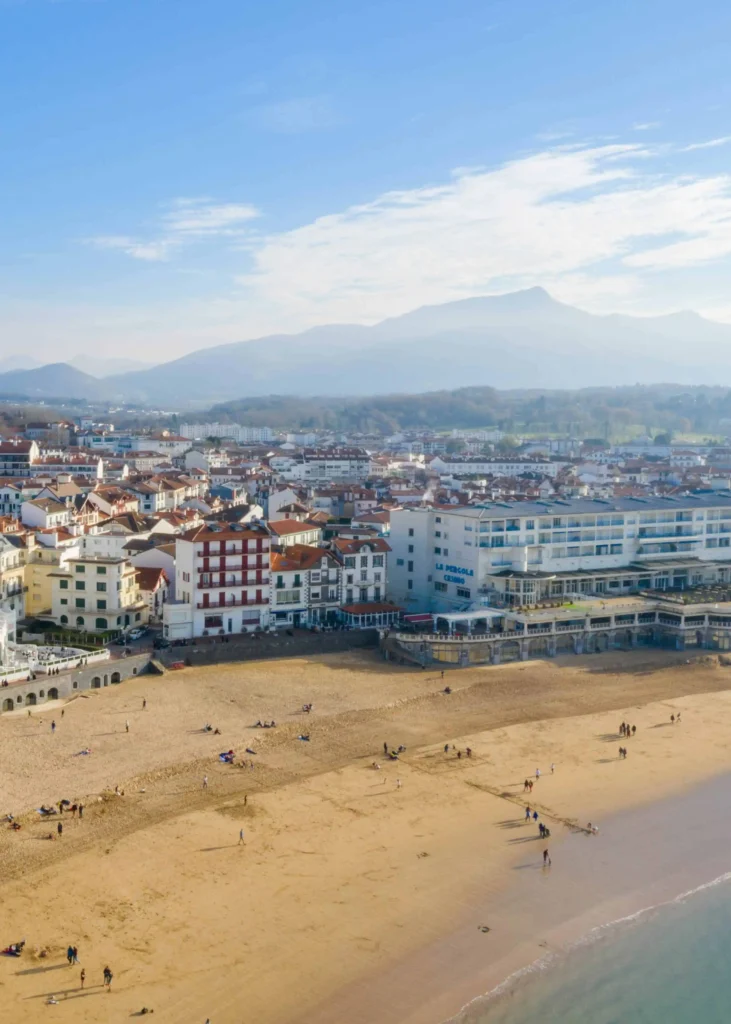 A panoramic view of a coastal town in the South of France, with sandy beaches, traditional buildings, and distant mountains.