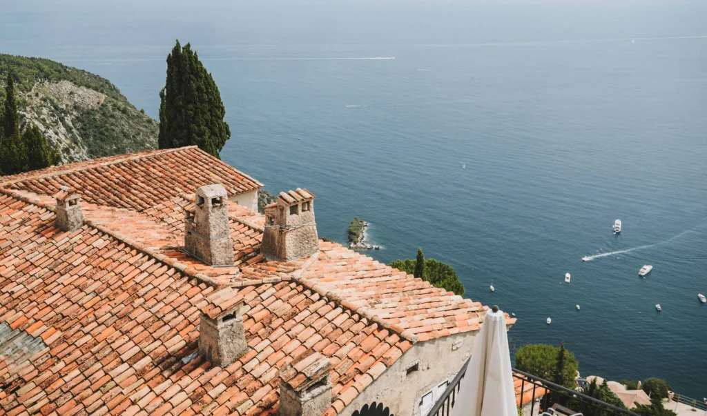 A rooftop view of terracotta-tiled houses perched on a hillside, with a stunning azure sea stretching into the horizon, dotted with small boats."