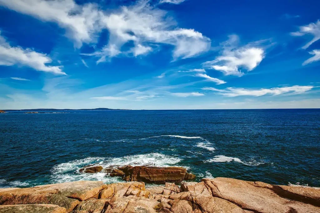 A rocky shoreline in Maine with waves crashing against the rocks, under a vibrant blue sky with scattered white clouds.