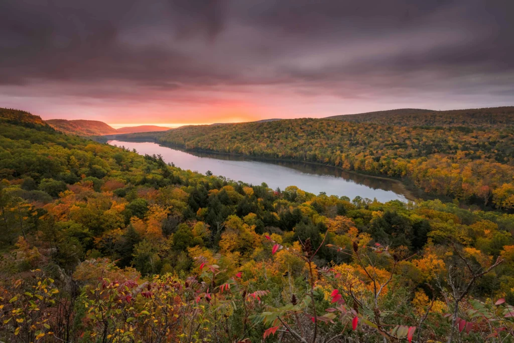 A stunning sunset view over Lake of the Clouds in Michigan, with vibrant orange and pink skies reflecting on the water, surrounded by a forest in peak autumn colors.