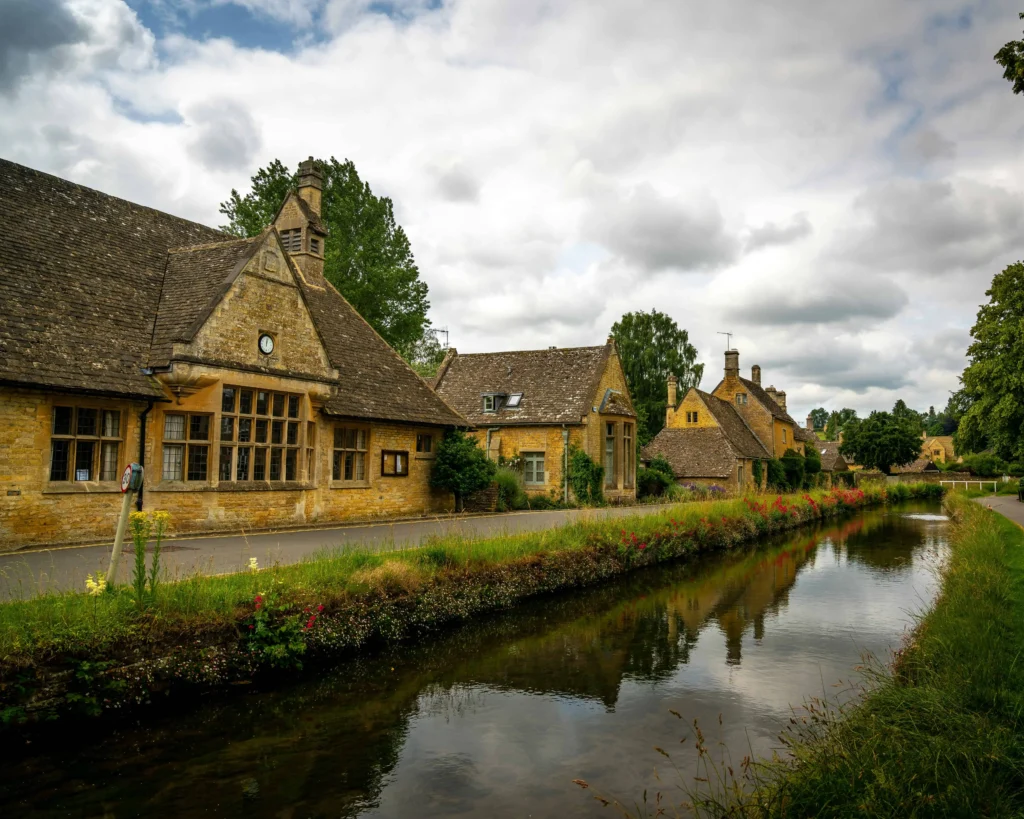 A picturesque Cotswolds village with stone cottages, a tranquil canal, and lush greenery under a cloudy sky.