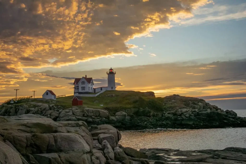 A coastal lighthouse on a rocky hill surrounded by dramatic clouds and golden hues of a sunset, highlighting the tranquil beauty of Maine's coastline.