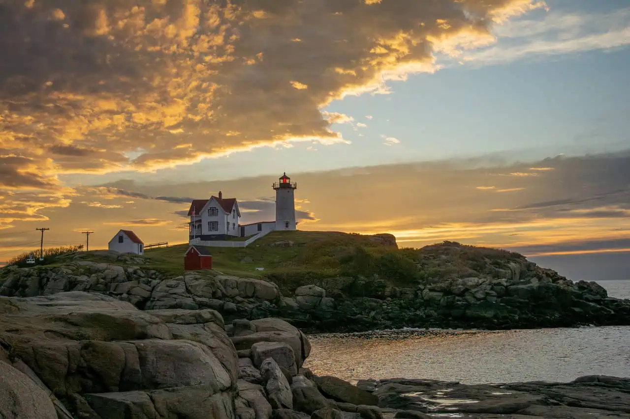 A coastal lighthouse on a rocky hill surrounded by dramatic clouds and golden hues of a sunset, highlighting the tranquil beauty of Maine's coastline.