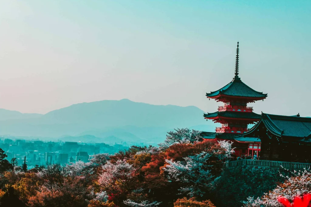 A traditional Japanese temple perched on a hill, with Kyoto’s cityscape and mountains in the background during cherry blossom season.