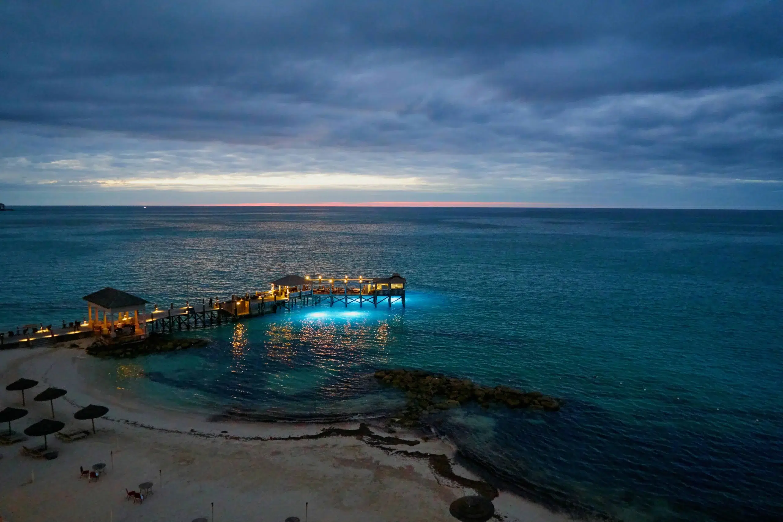 A serene beach in Nassau at dusk, featuring an illuminated pier and calm ocean waves under a cloudy sky.