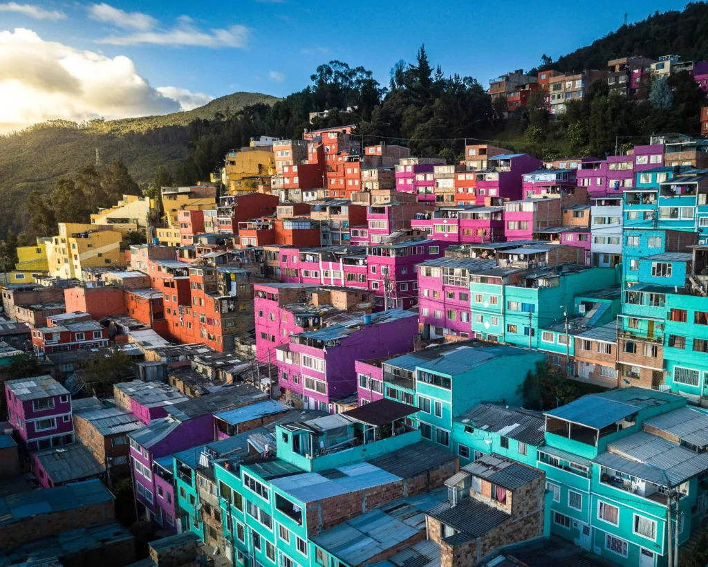 A vibrant hillside neighborhood in Colombia featuring colorful houses in shades of pink, blue, and yellow, surrounded by lush greenery and a clear blue sky