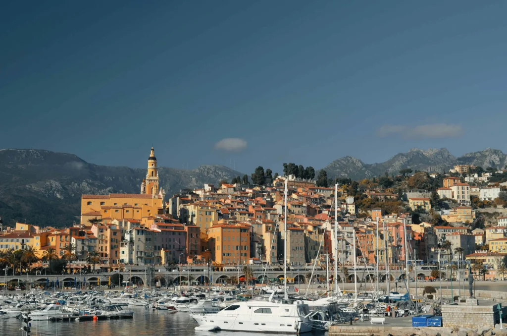 A daytime view of the vibrant town of Menton, France, with its orange and pastel-colored buildings set against a mountainous backdrop and a harbor filled with yachts