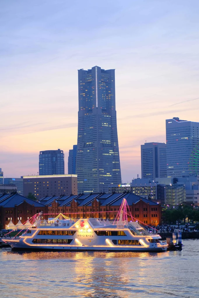 Yokohama’s Landmark Tower at sunset with a brightly lit boat and the Red Brick Warehouse in the foreground.