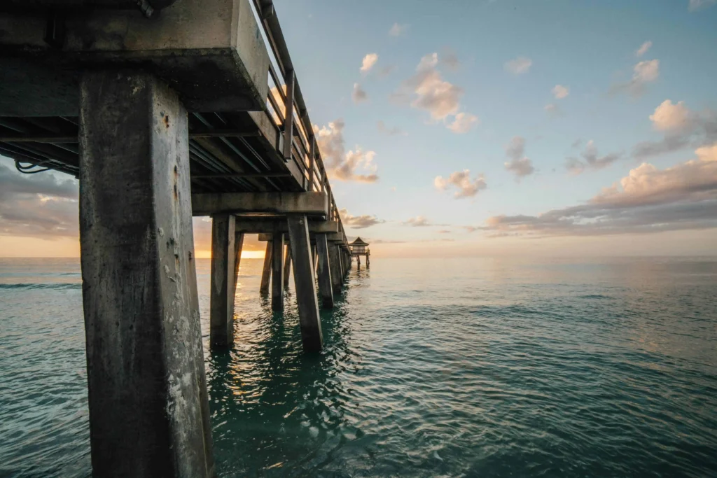 A striking perspective of Naples Pier from underneath, highlighting the weathered wooden supports and shimmering blue ocean waters.
