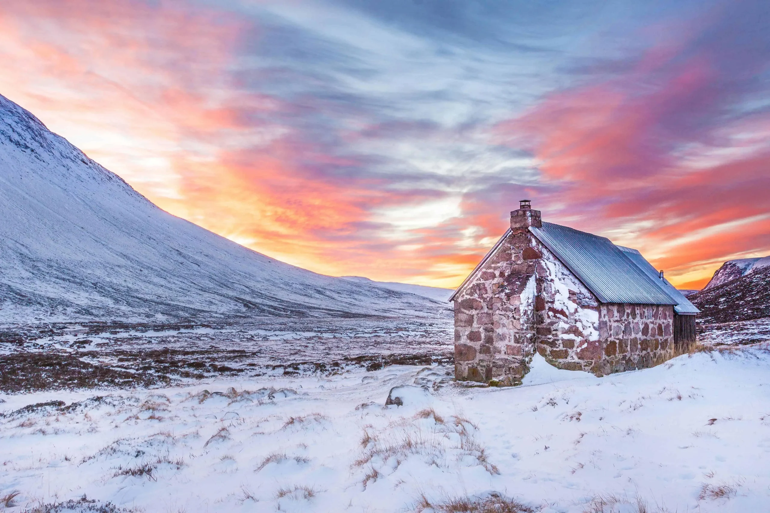 A cozy stone cottage in a snow-covered Scottish landscape with a vibrant sunrise casting pink and orange hues across the sky.