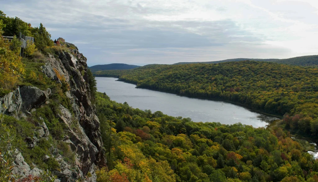 A dramatic view from a cliff overlooking Lake of the Clouds in Michigan, surrounded by a lush green forest on a partly cloudy day.