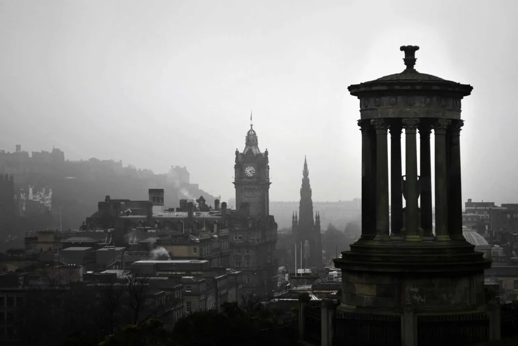 A misty view of Edinburgh, Scotland, showcasing the iconic Balmoral Clock Tower, Scott Monument, and a historic skyline from Calton Hill.