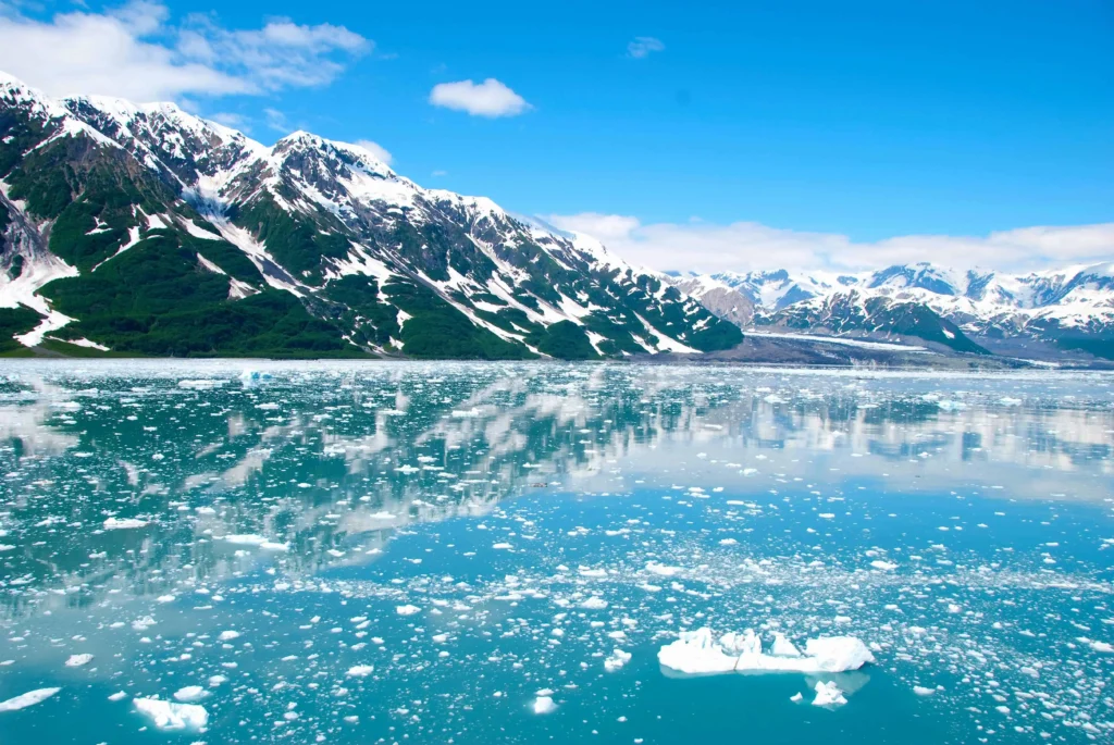 A serene view of snow-covered mountains and floating ice in a turquoise glacial lake under a clear blue sky in Alaska