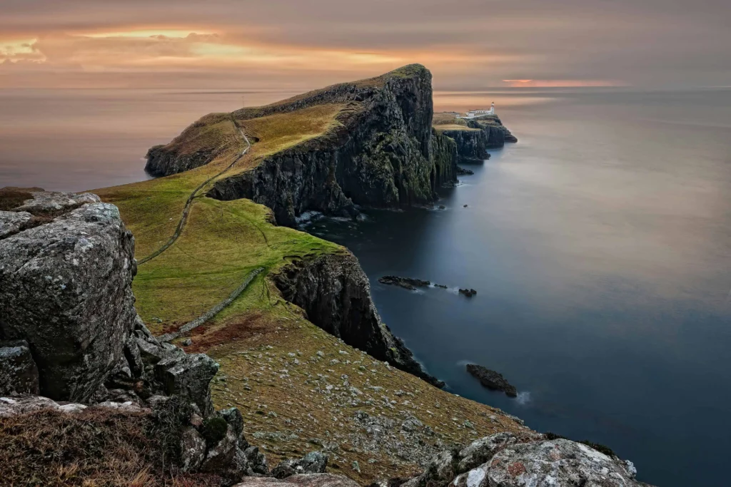 Neist Point Lighthouse in Scotland, situated on a rugged cliff overlooking the calm sea during a serene sunset.