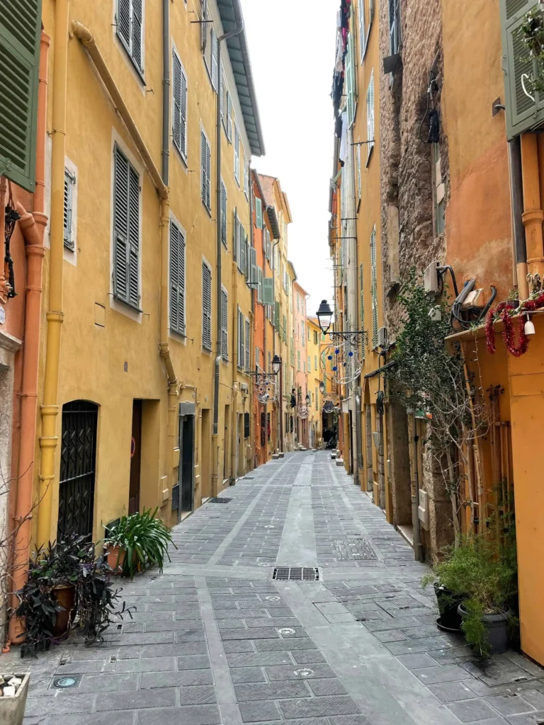 A narrow, picturesque street in Menton, lined with yellow and orange houses, shuttered windows, and plants in pots along the pathway.