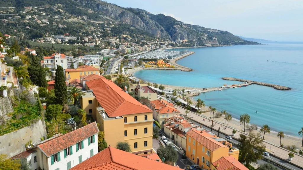 A panoramic view of Menton’s coastline, with red-roofed houses and a clear turquoise sea bordered by green hills and rocky outcrops.