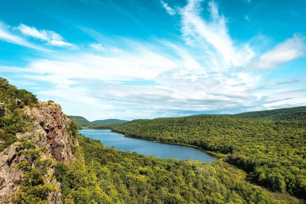 A clear and bright summer day at Lake of the Clouds in Michigan, featuring a panoramic view of the blue waters and green forested hills beneath a sky streaked with white clouds.