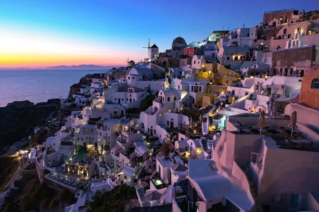 Sunset view over Oia, Santorini, with illuminated whitewashed buildings and windmills, creating a romantic atmosphere ideal for a honeymoon in Greece.