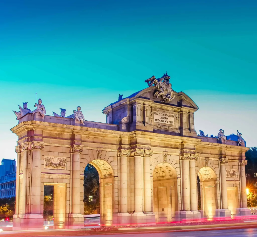 The Puerta de Alcalá monument in Madrid, Spain, lit up against a vibrant evening sky.