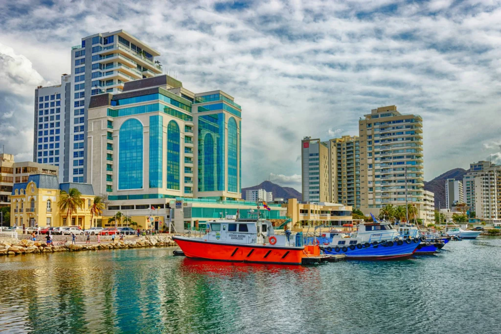 vibrant marina in Santa Marta, Colombia, featuring modern high-rise buildings with glass facades, colorful fishing boats, and a scenic backdrop of mountains under a partly cloudy sky.