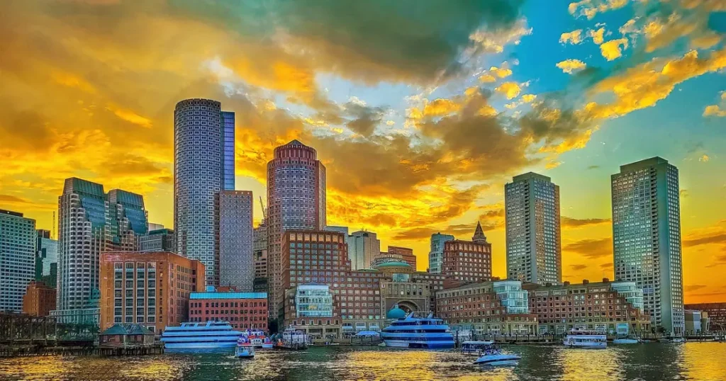 Stunning view of the Boston skyline during sunset with vibrant orange and yellow hues reflecting on the buildings and water.