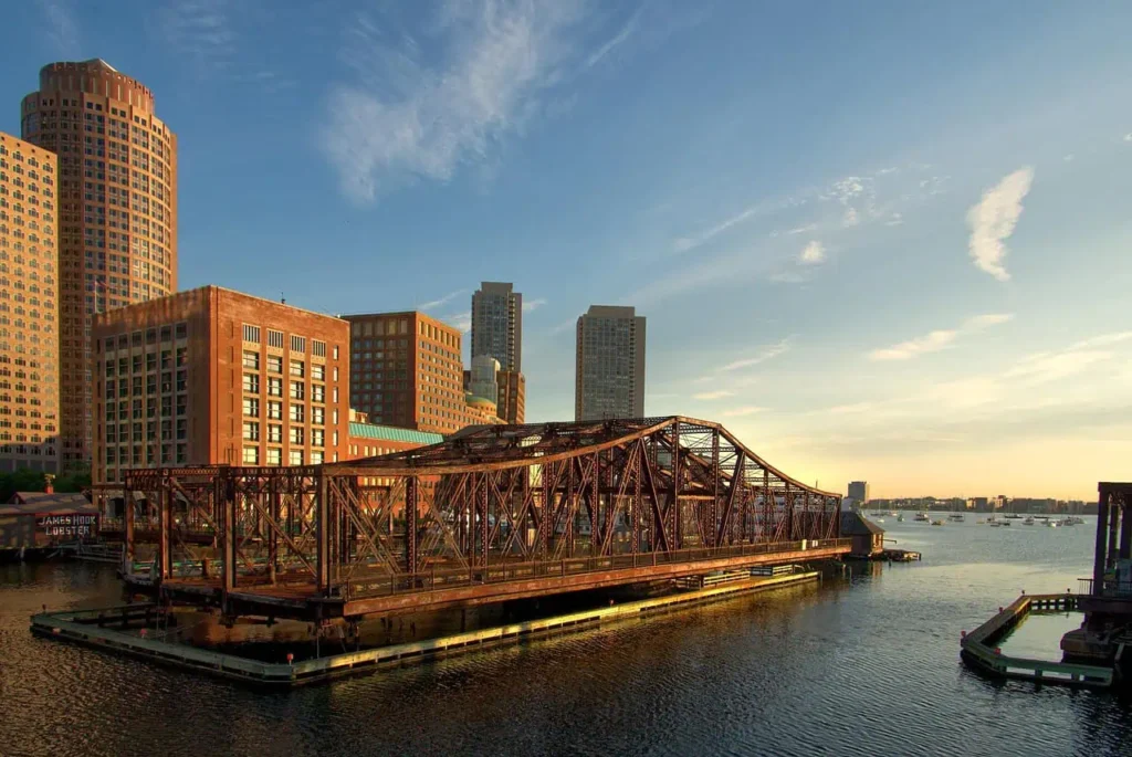 A rusted bridge structure in Boston Harbor with city buildings in the background during golden hour.