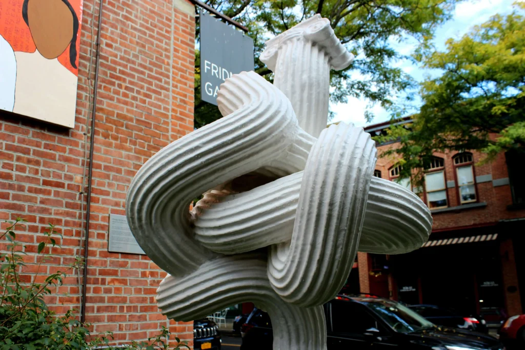 A white abstract sculpture resembling intertwined columns set in a city garden.