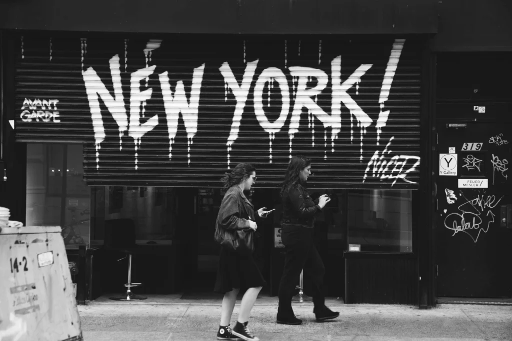 Two women walking past a graffiti-covered storefront with "New York!" prominently displayed.