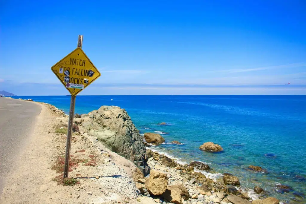 A coastal road with a sign warning of falling rocks and vibrant blue ocean in Catalina Island.