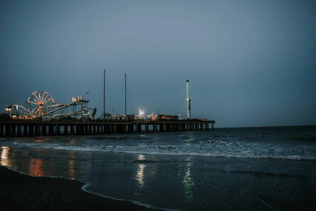 A glowing pier at night with a Ferris wheel and other amusement rides reflected on the water