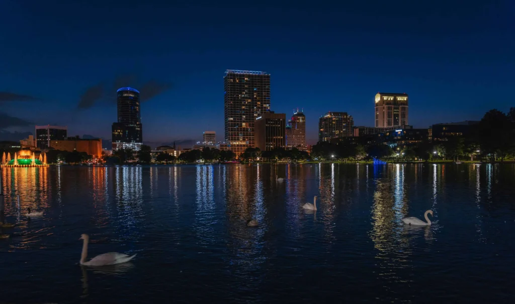 A serene nighttime view of Lake Eola Park in Orlando, featuring city skyline reflections on the water and swans gliding across the lake.