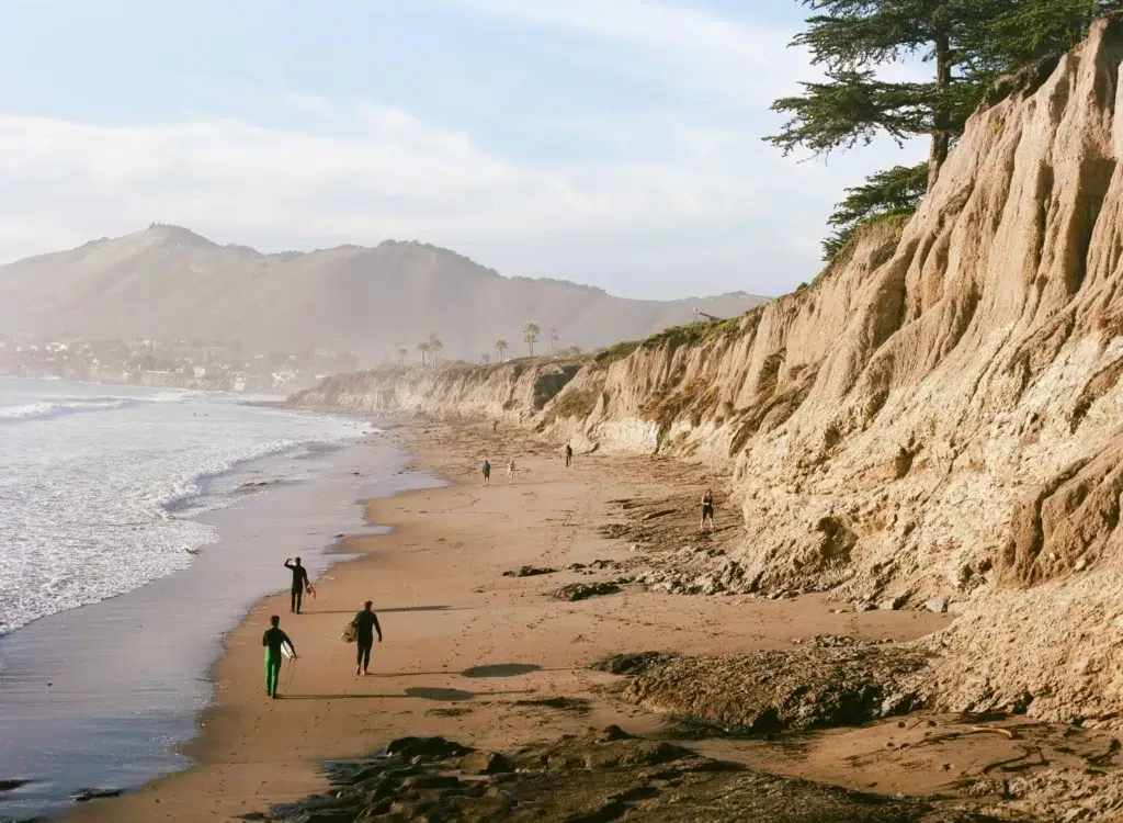 Beachgoers and surfers walking along the sandy shoreline with cliffs and ocean waves in the background.