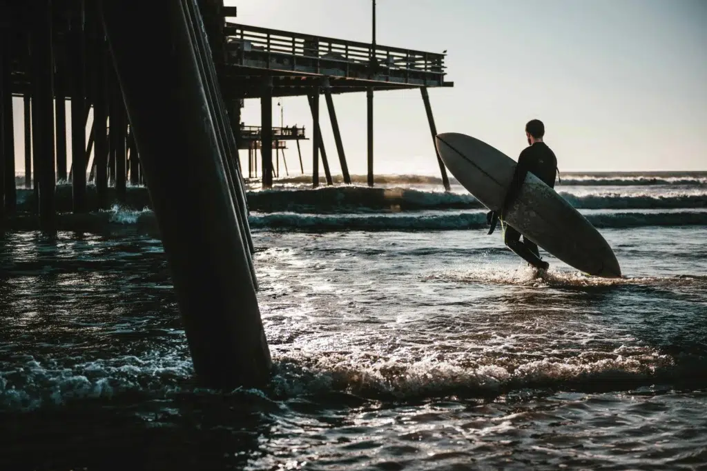 A lone surfer holding a board, walking under the Pismo Beach pier at sunset.