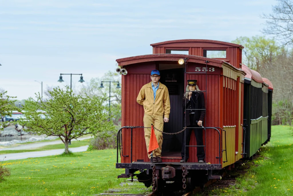 A vintage red train with two conductors standing on the back platform in Portland, Maine.