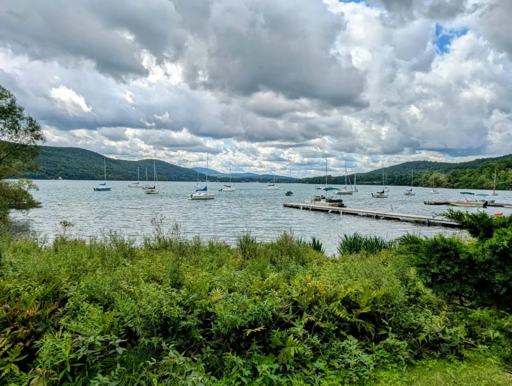 A lakefront view with sailboats on Otsego Lake in Cooperstown, NY, under a cloudy sky.